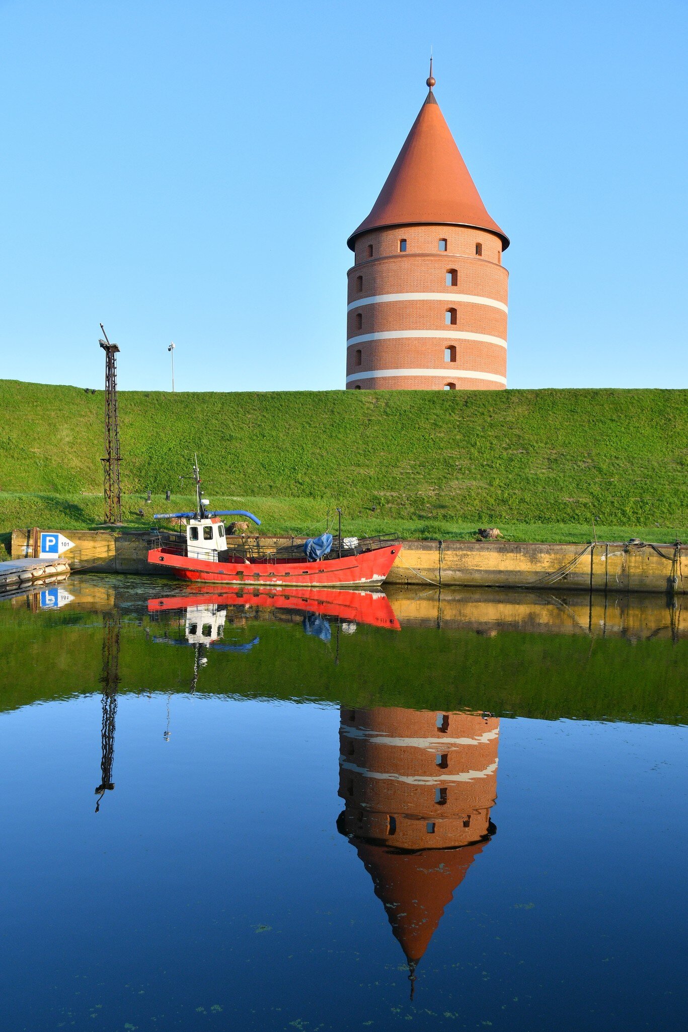 A round red-brick tower with white bands and a conical tiled roof standing on a grass rampart, mirrored in still water with a red fishing boat moored below.