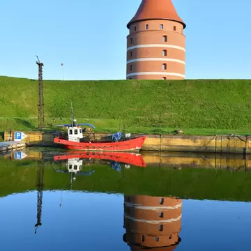A round red-brick tower with white bands and a conical tiled roof standing on a grass rampart, mirrored in still water with a red fishing boat moored below.