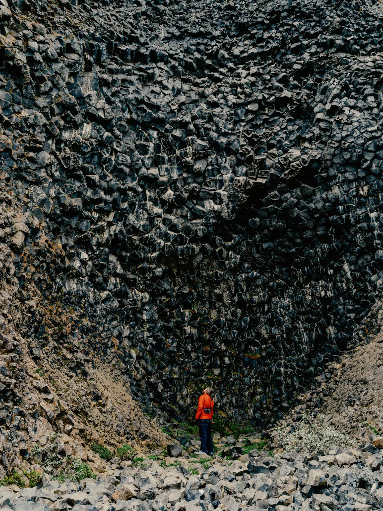 A man in an orange jacket stands at the bottom of a rocky outcropping in Iceland