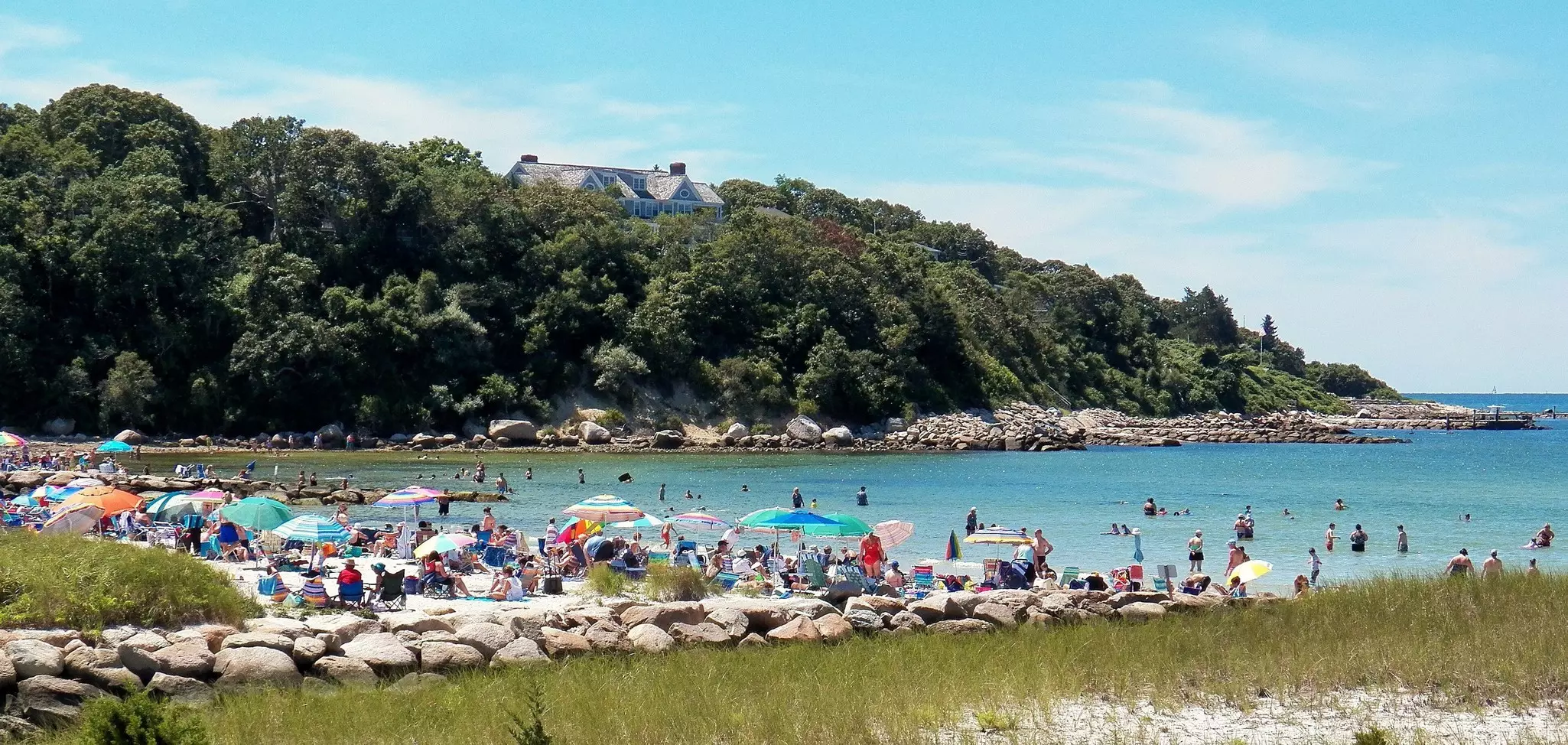 A beach lined with colorful sunshades and crowded with sunbathers and swimmers in the ocean beyond on a sunny day. A large-roofed home peeks through a forested hillside in the distance.