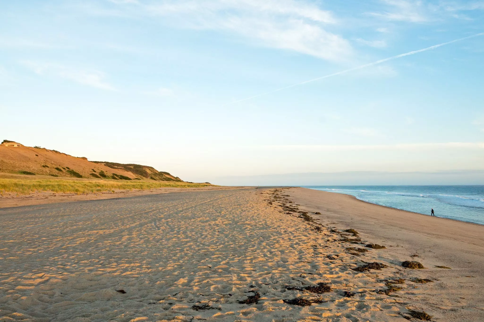 Sunrise along a wide, sandy beach with dunes to the left and the ocean to the right. One person walks along the short in the distance.