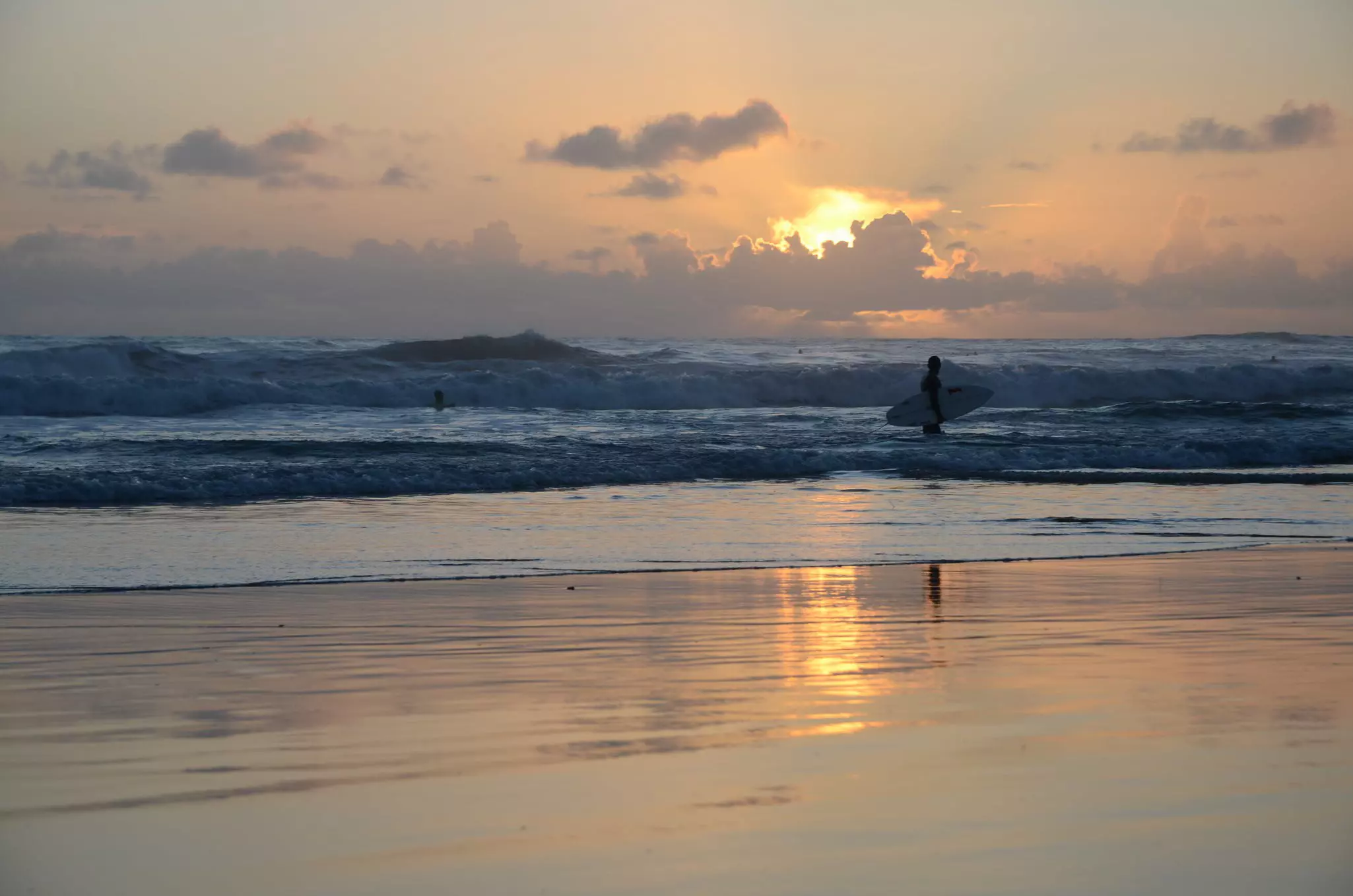Surfer carrying surfboard near cresting waves with setting sun reflected on the sand.