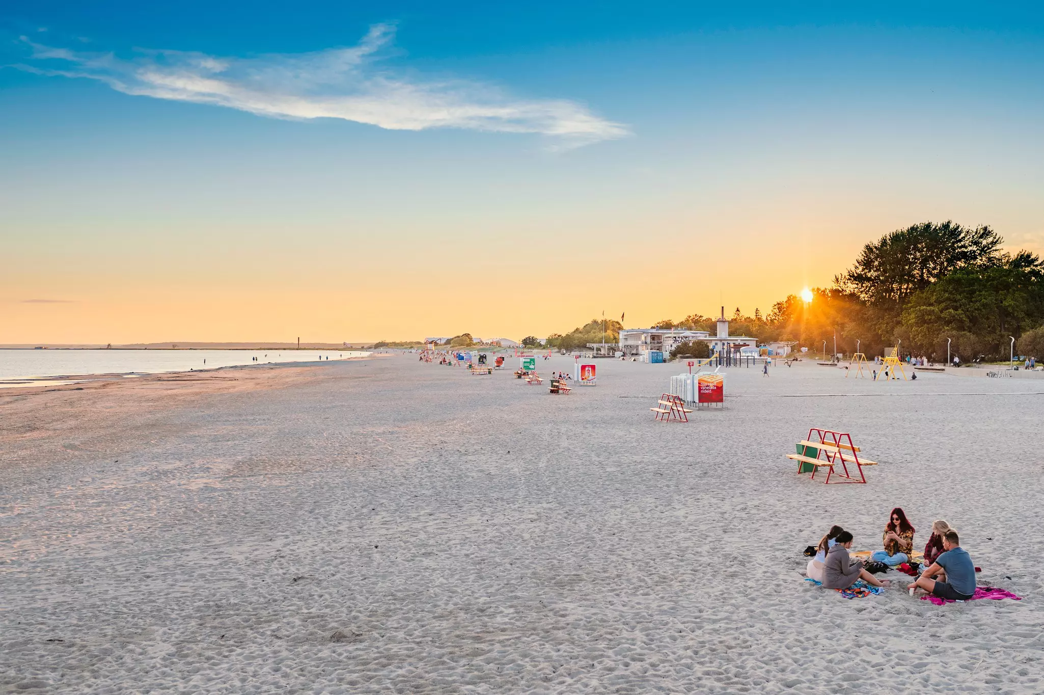 A wide sandy beach with people sat in small groups enjoying the last light of a summer's day. Some people are paddling in the shallows.