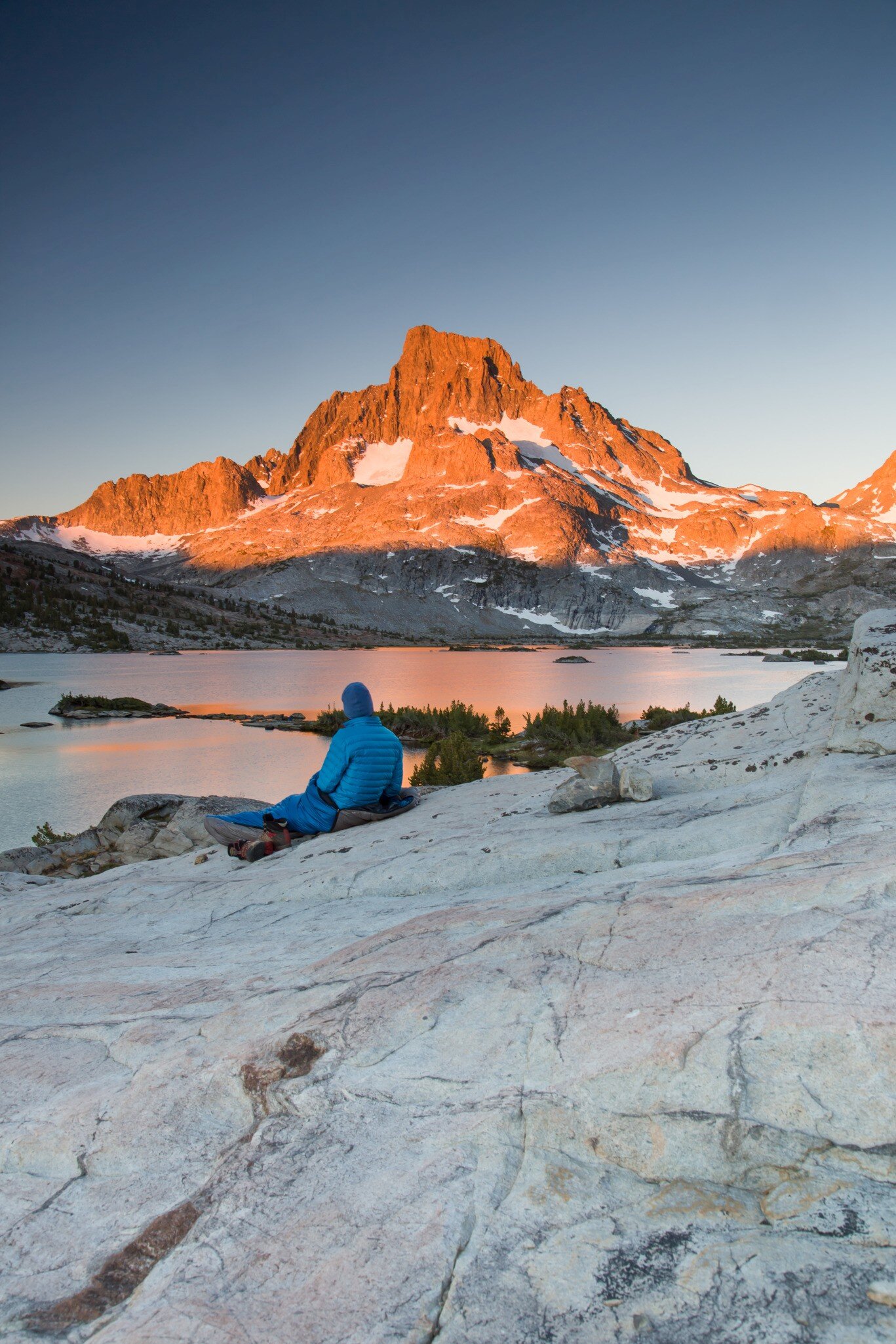 A lone backpacker with a blue stocking cap sits on a slab of granite rock and watches the early sun beams of the morning light hit Banner Peak behind Thousand Island Lake in the Ansel Adams Wilderness.