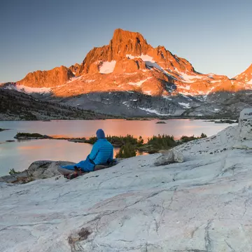 A lone backpacker with a blue stocking cap sits on a slab of granite rock and watches the early sun beams of the morning light hit Banner Peak behind Thousand Island Lake in the Ansel Adams Wilderness.