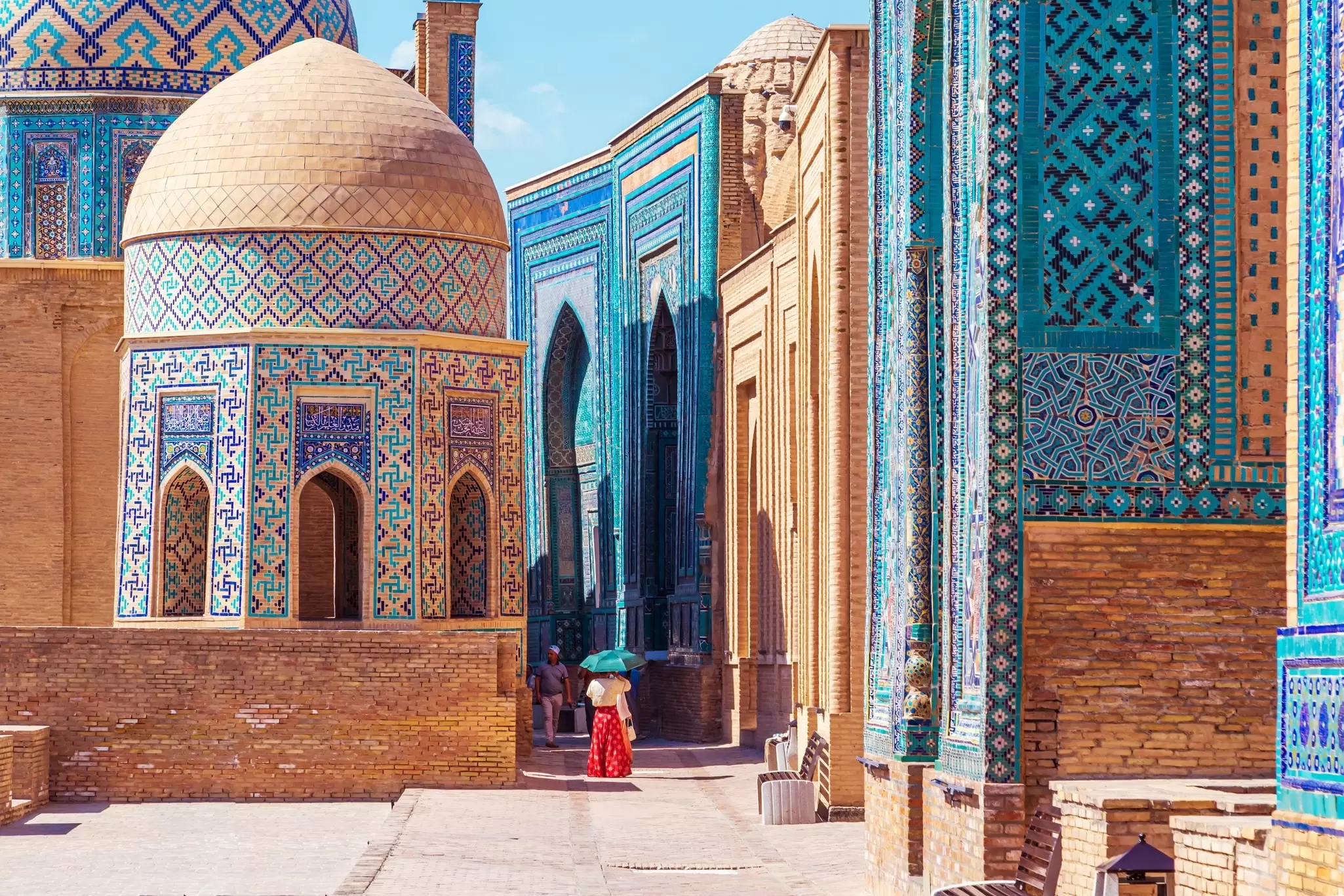 A person in red walks toward buildings covered with turquoise and blue tiles in geographic patterns.