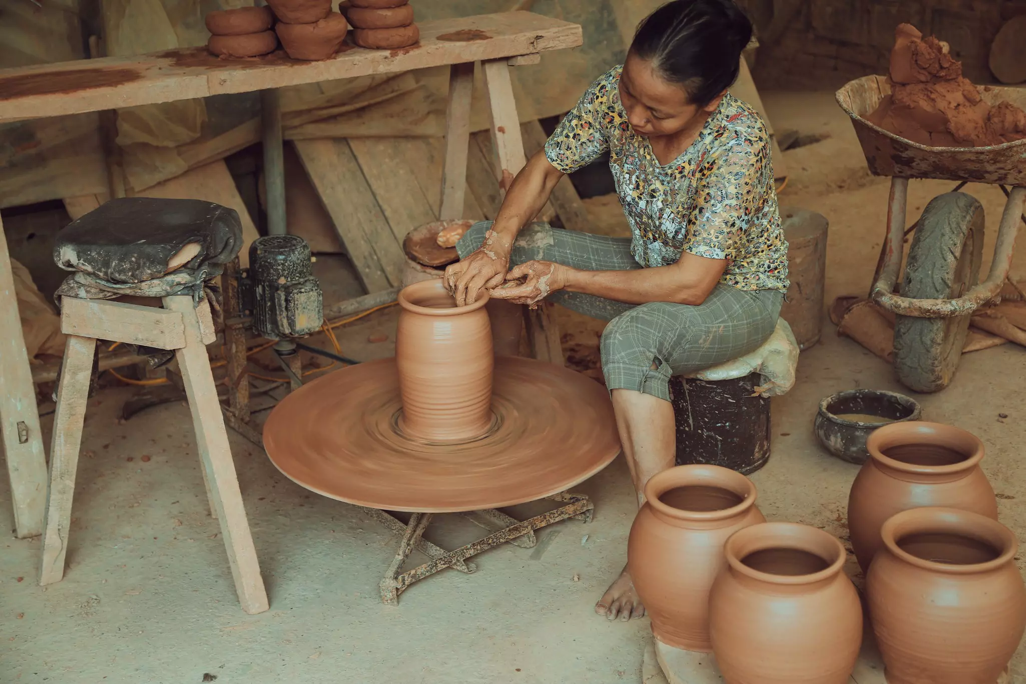 A woman sits and works at a potter's wheel, surrounded by vases
