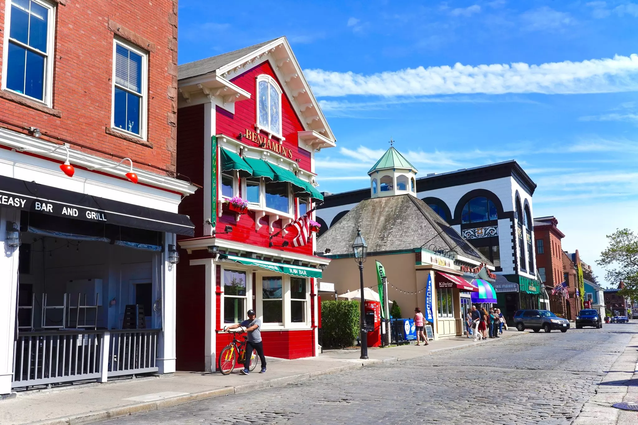 Cobblestone street bordered by brick and red-shingled buildings on a sunny day, with pedestrians and a man pushing a bicycle on the sidewalk.