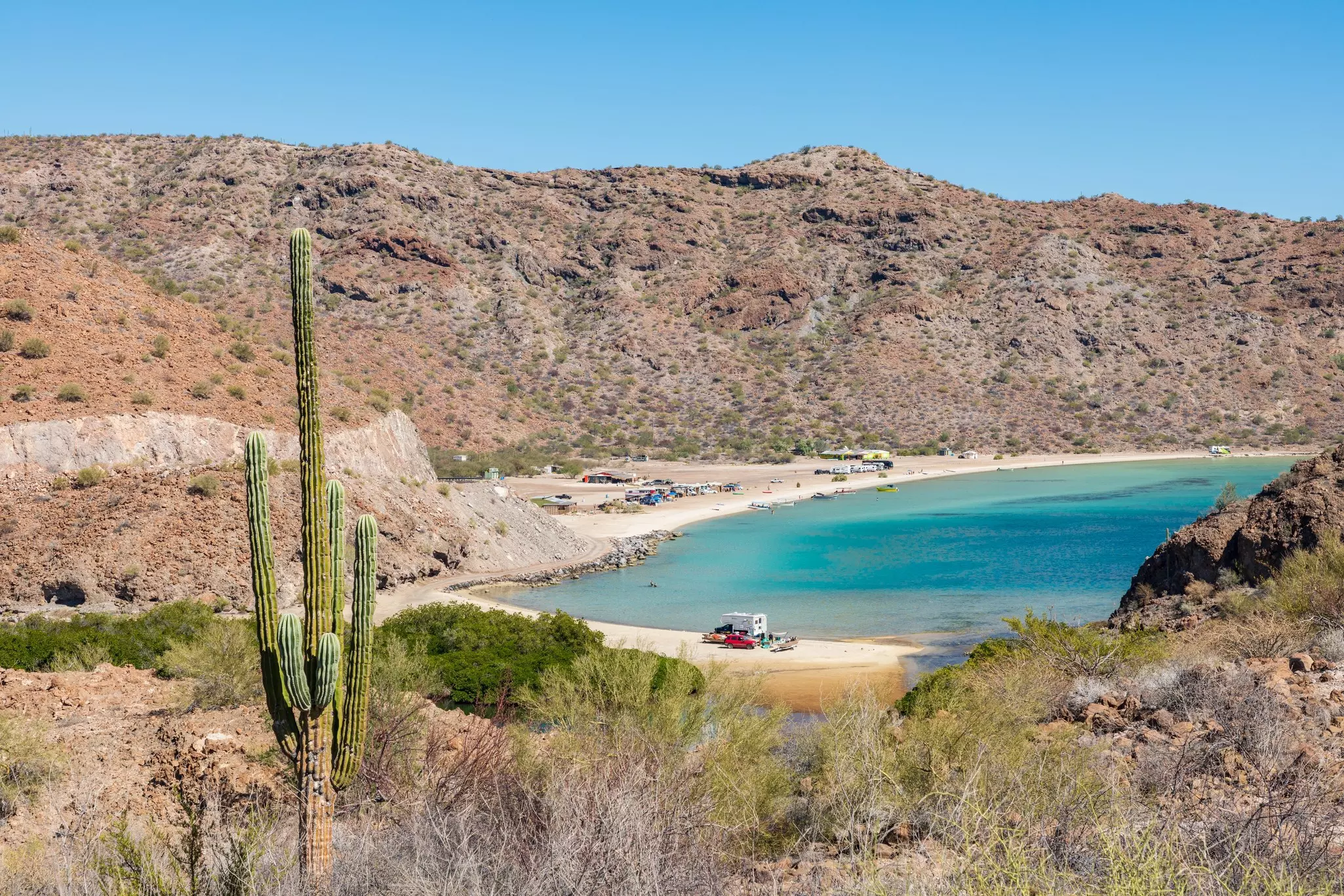Santispac beach on the Baja peninsula, Mexico with a cactus in the foreground and cars on the beach in the centre.