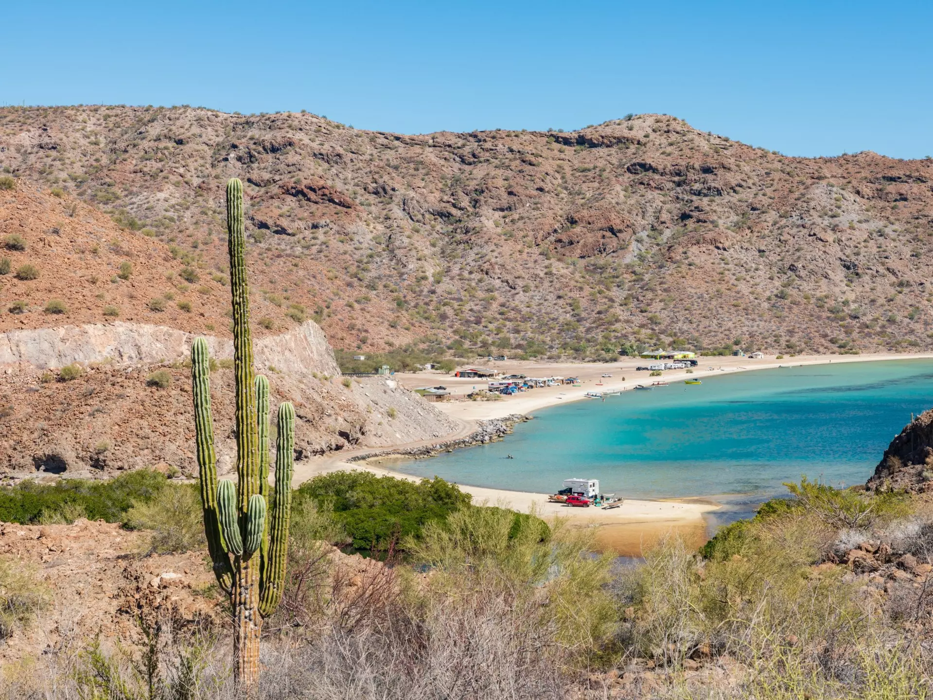Santispac beach on the Baja peninsula, Mexico with a cactus in the foreground and cars on the beach in the centre.