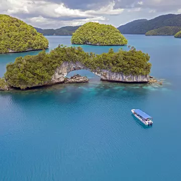 A small boat travels through dark blue ocean towards an island with a large rock arch.