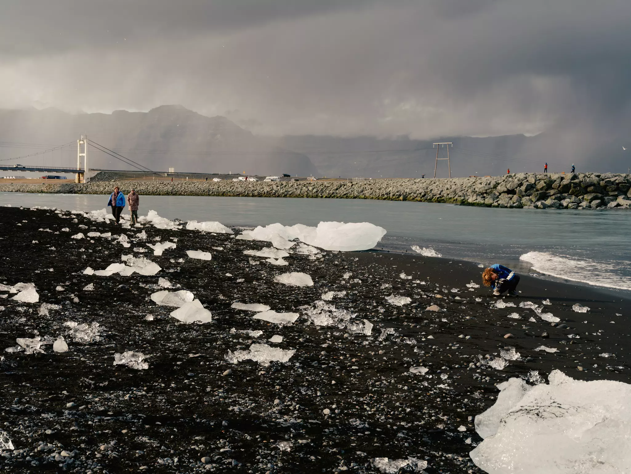 People walk on a black sand beach with large chunks of clear ice.