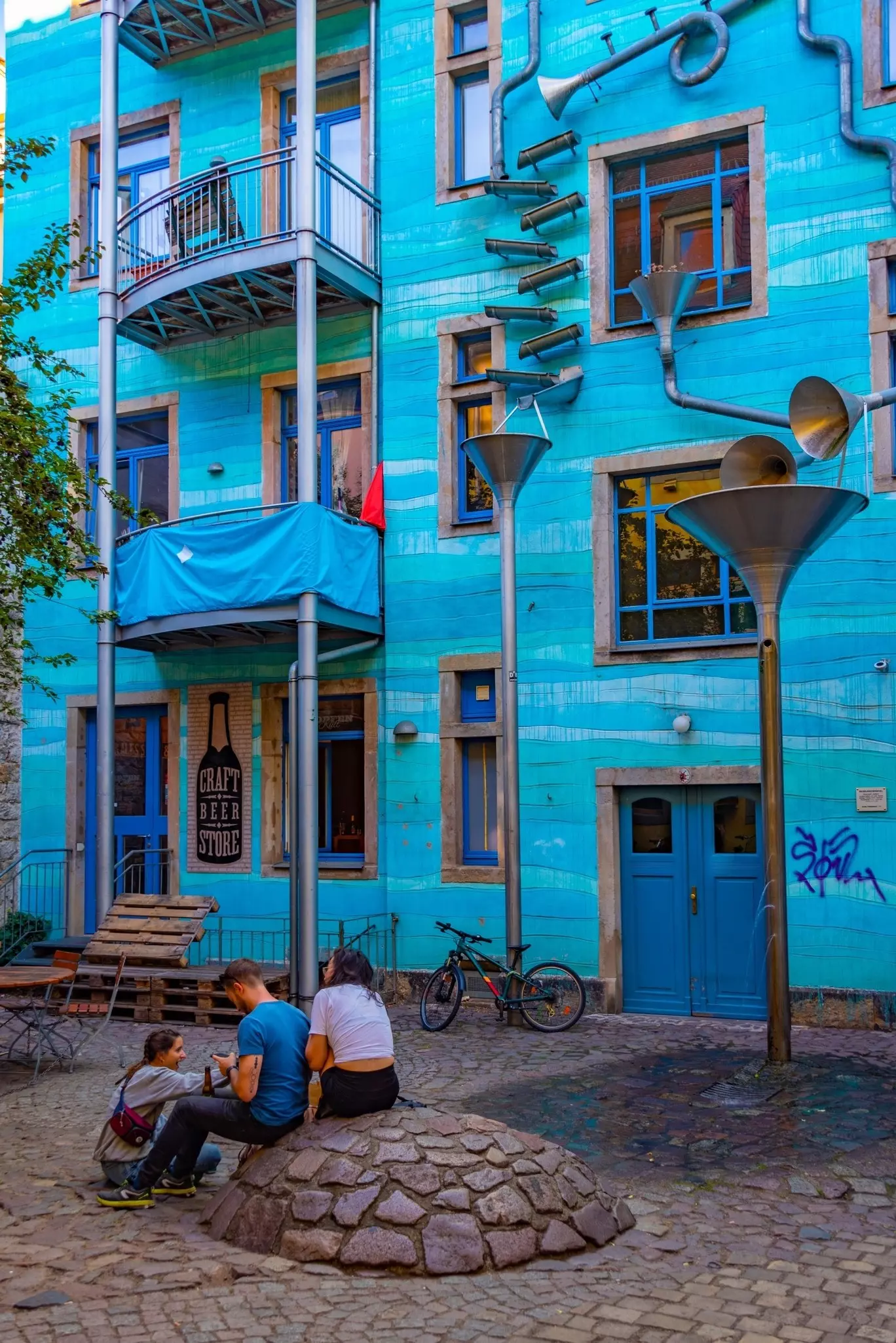 Three young people sit on the ground chatting outside a tall blue residential building with a craft beer shop on the ground floor.