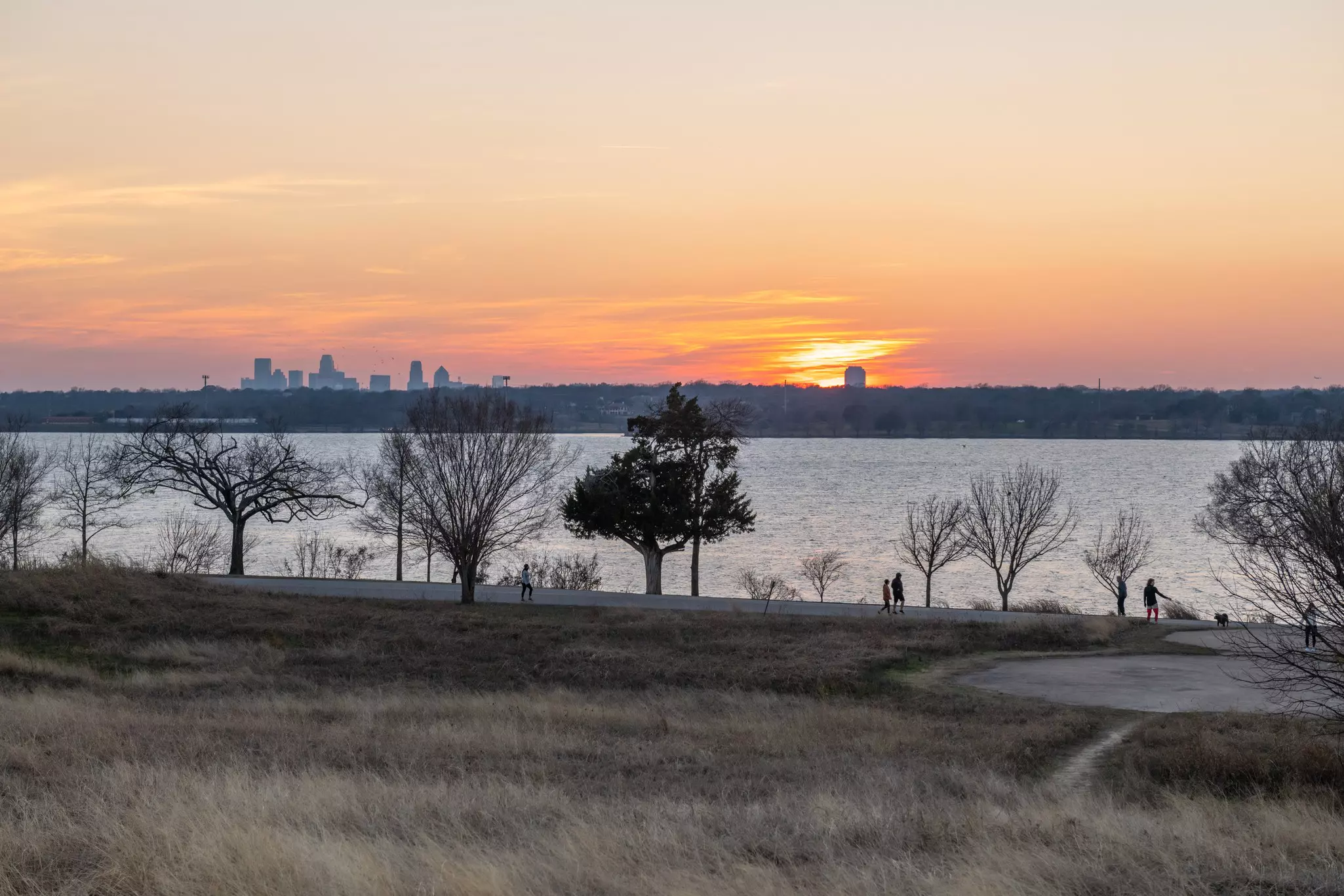 People walking along a path at sunset overlooking a lake with a city skyline in the distance.