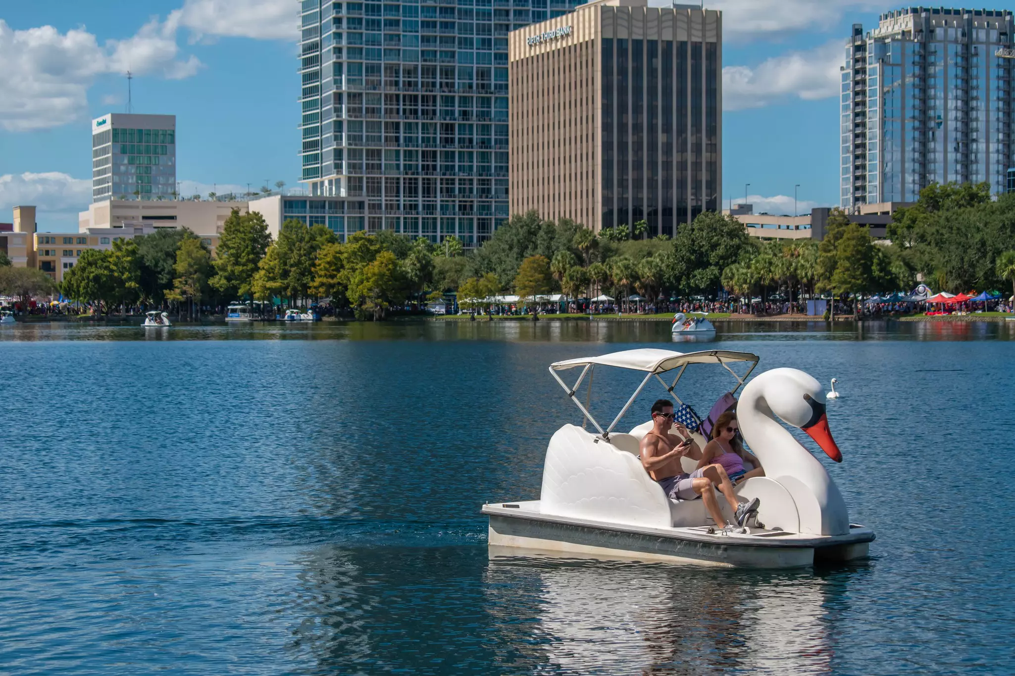 Two people use their feet to paddle a swan-shaped boat on a lake in a city, with a skyline of towers along the shore.