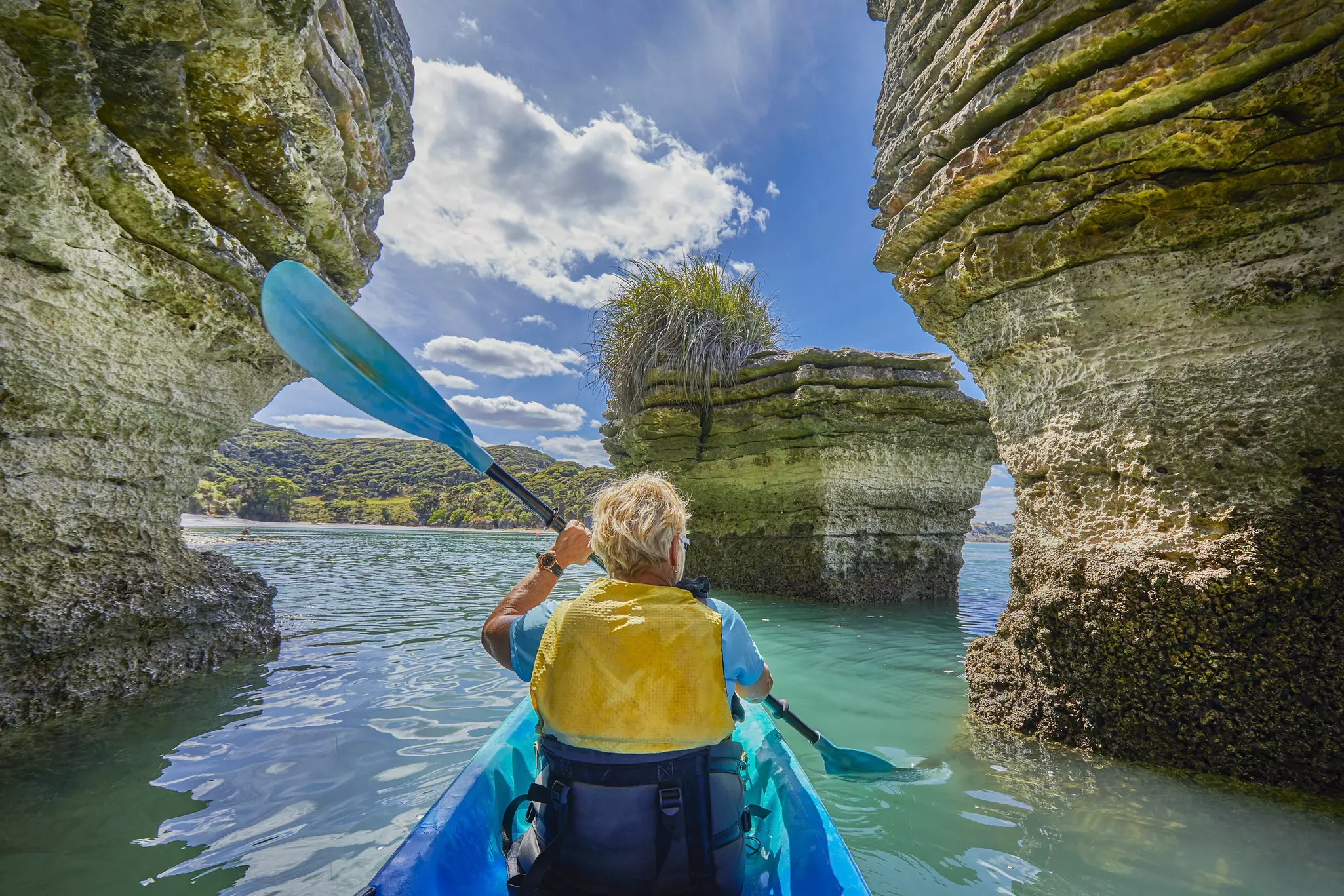 A person wearing a yellow life vest paddles a blue kayak toward rock formations in New Zealand.