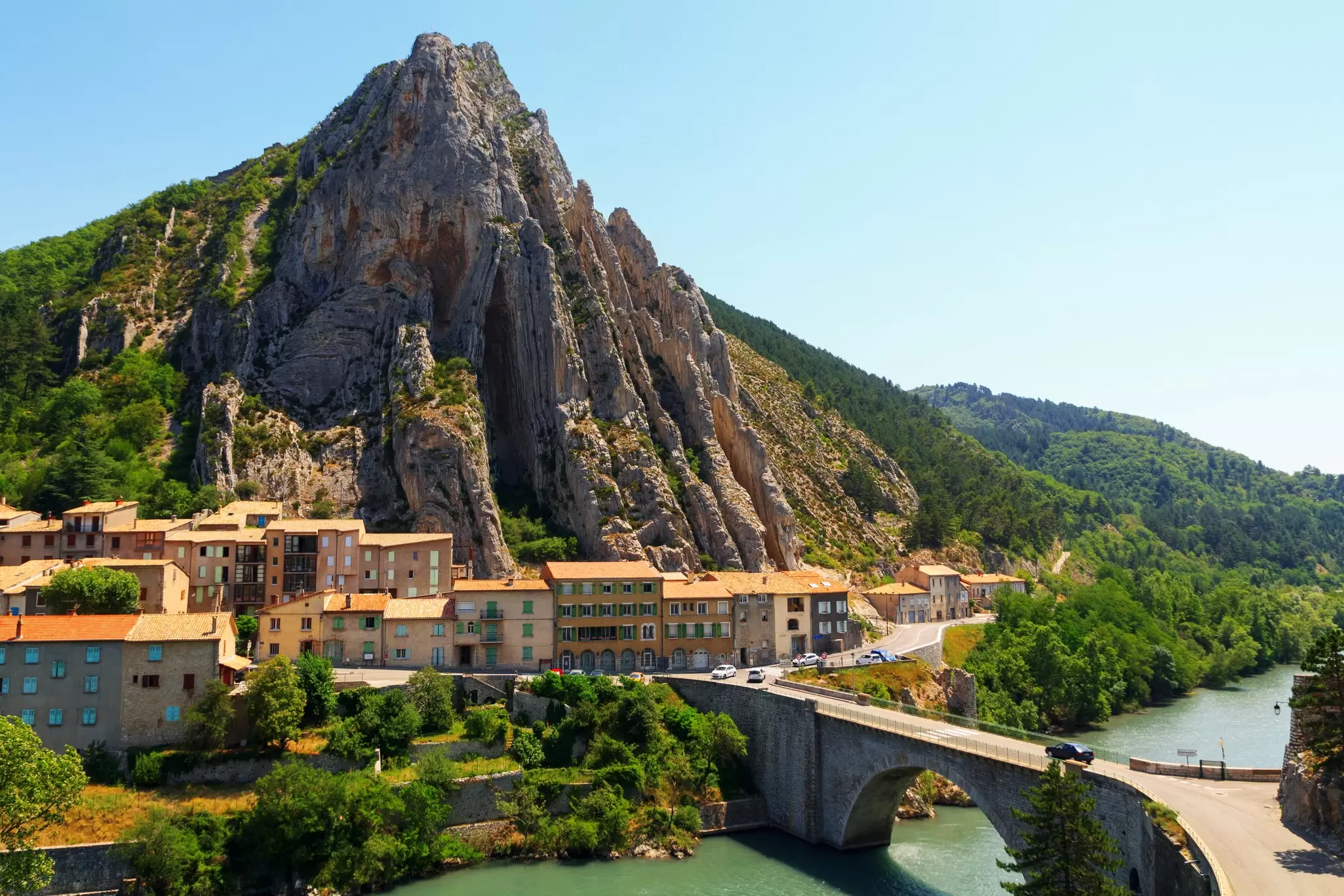 A town by a river across from a bridge is at the base of a large pointed rock formation.