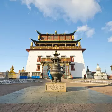 White multi-tiered temple with green-and-gold roofs behind a bronze urn on a plinth, a yellow hall and stupas nearby