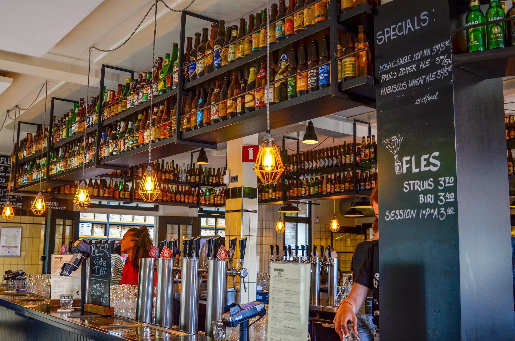 A bar counter with a line of taps. Beer bottles line shelves suspended from the ceiling.