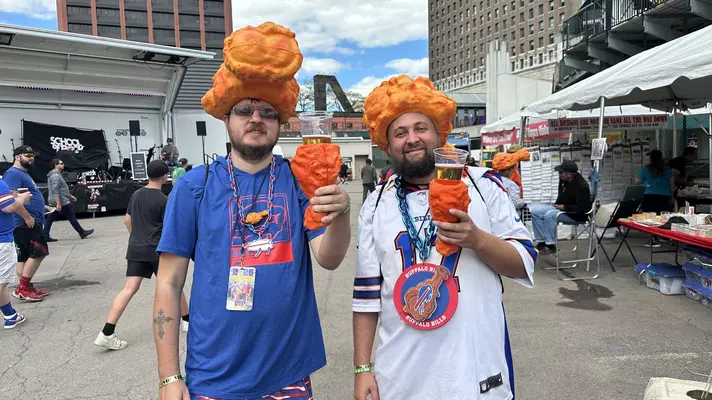 Two men in Buffalo Bills jerseys wearing foam chicken wing hats and chicken wing beer holders. 