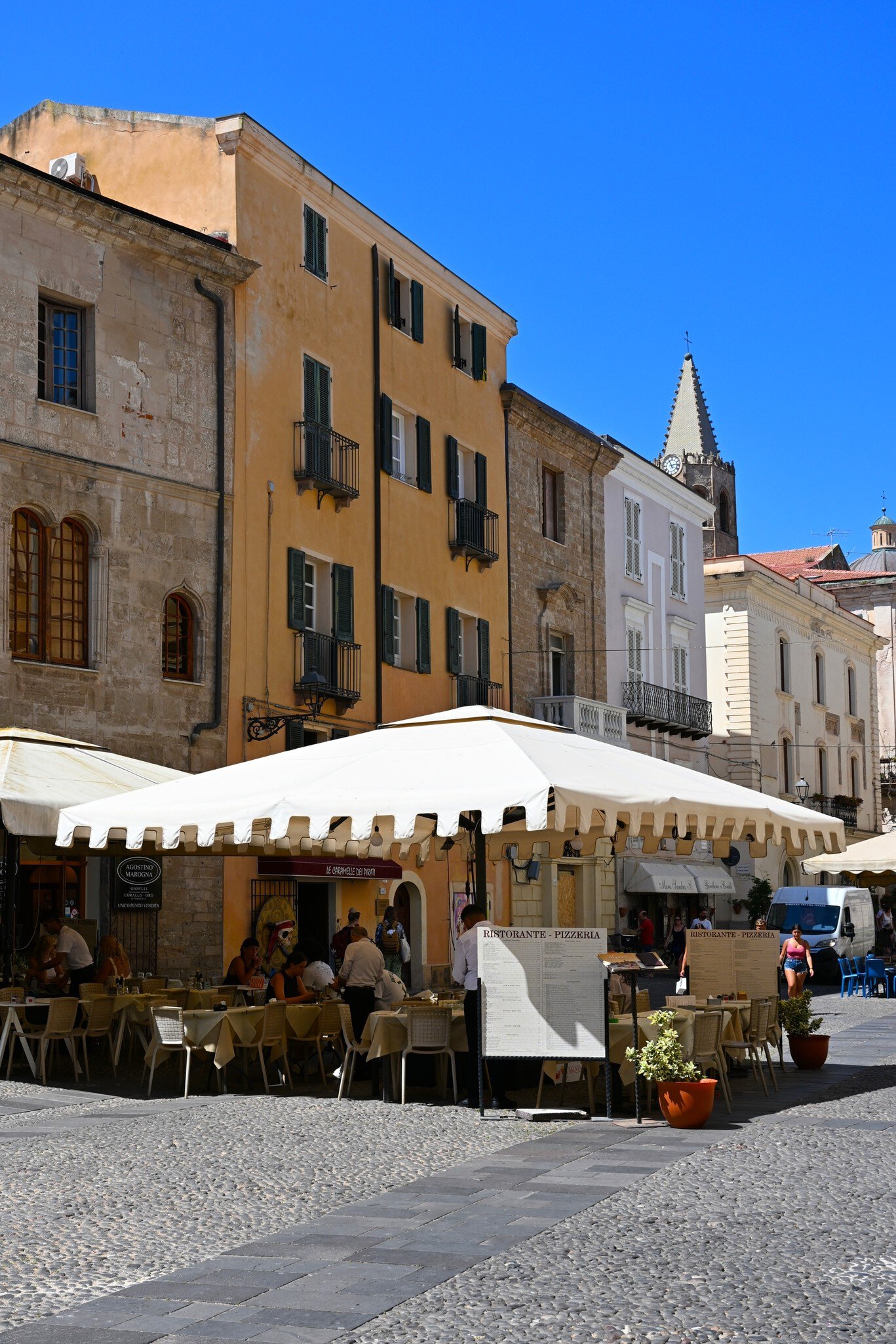 Outdoor restaurant under a white awning on a cobbled square in Alghero, with ochre buildings and a pointed bell tower.