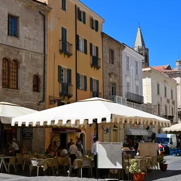Outdoor restaurant under a white awning on a cobbled square in Alghero, with ochre buildings and a pointed bell tower.