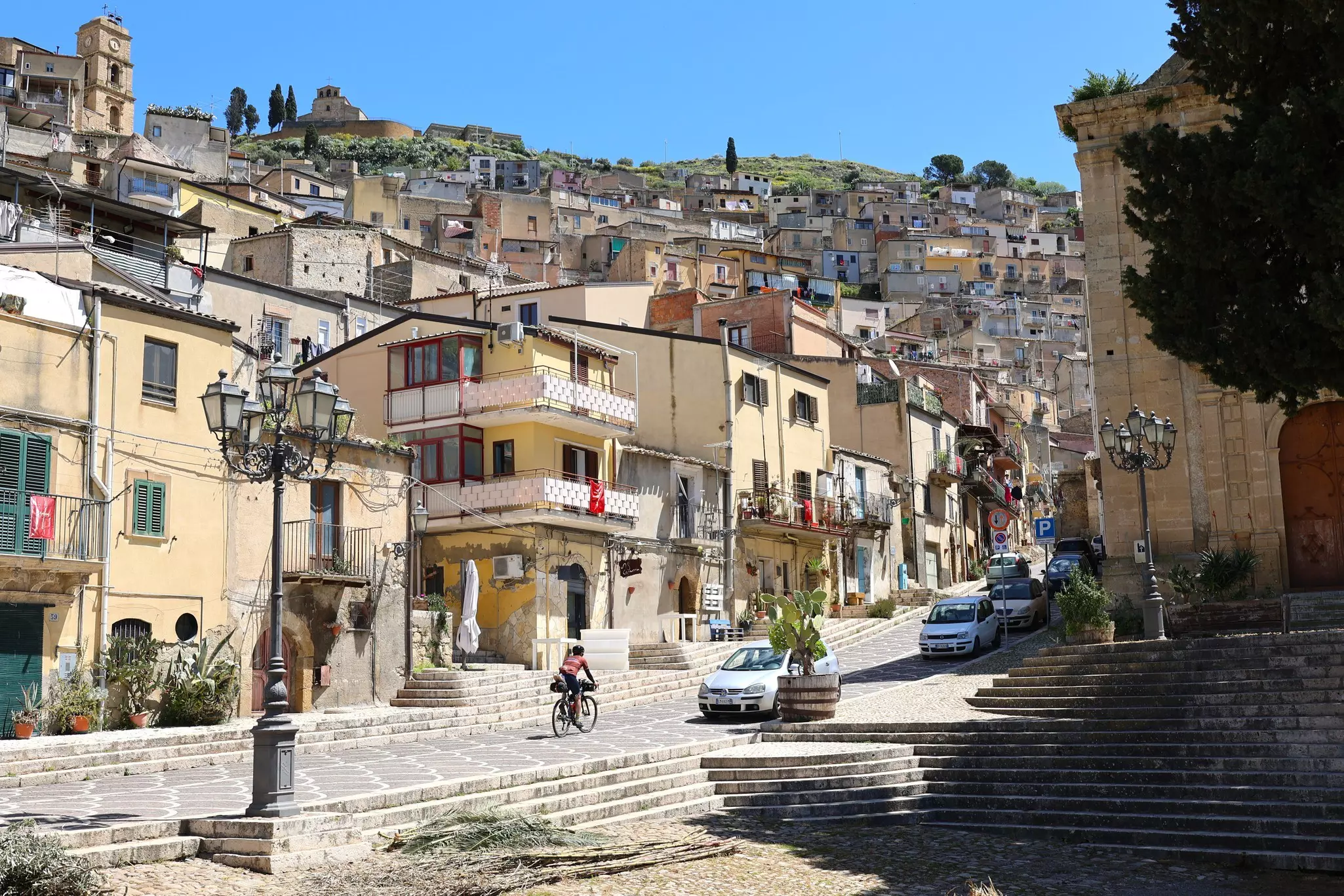 A cyclist pedals uphill on a quiet road through the center of a hilltop town.
