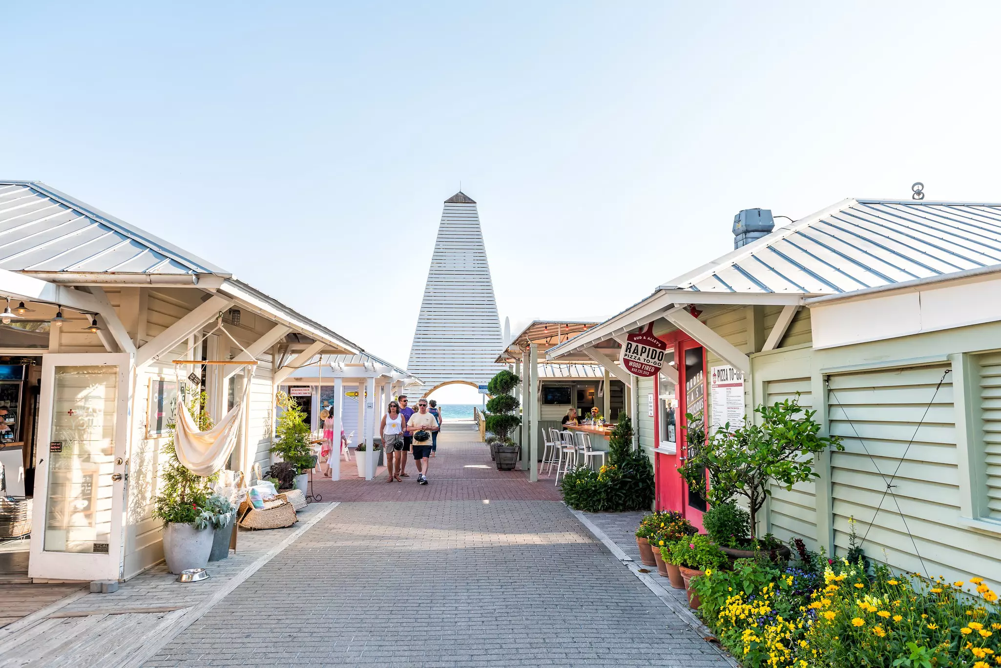 People walk through a pedestrian area in Florida with pastel-colored, one-story shops and cafes; a tower is at the end of the walkway, and the ocean is visible in the opening below.
