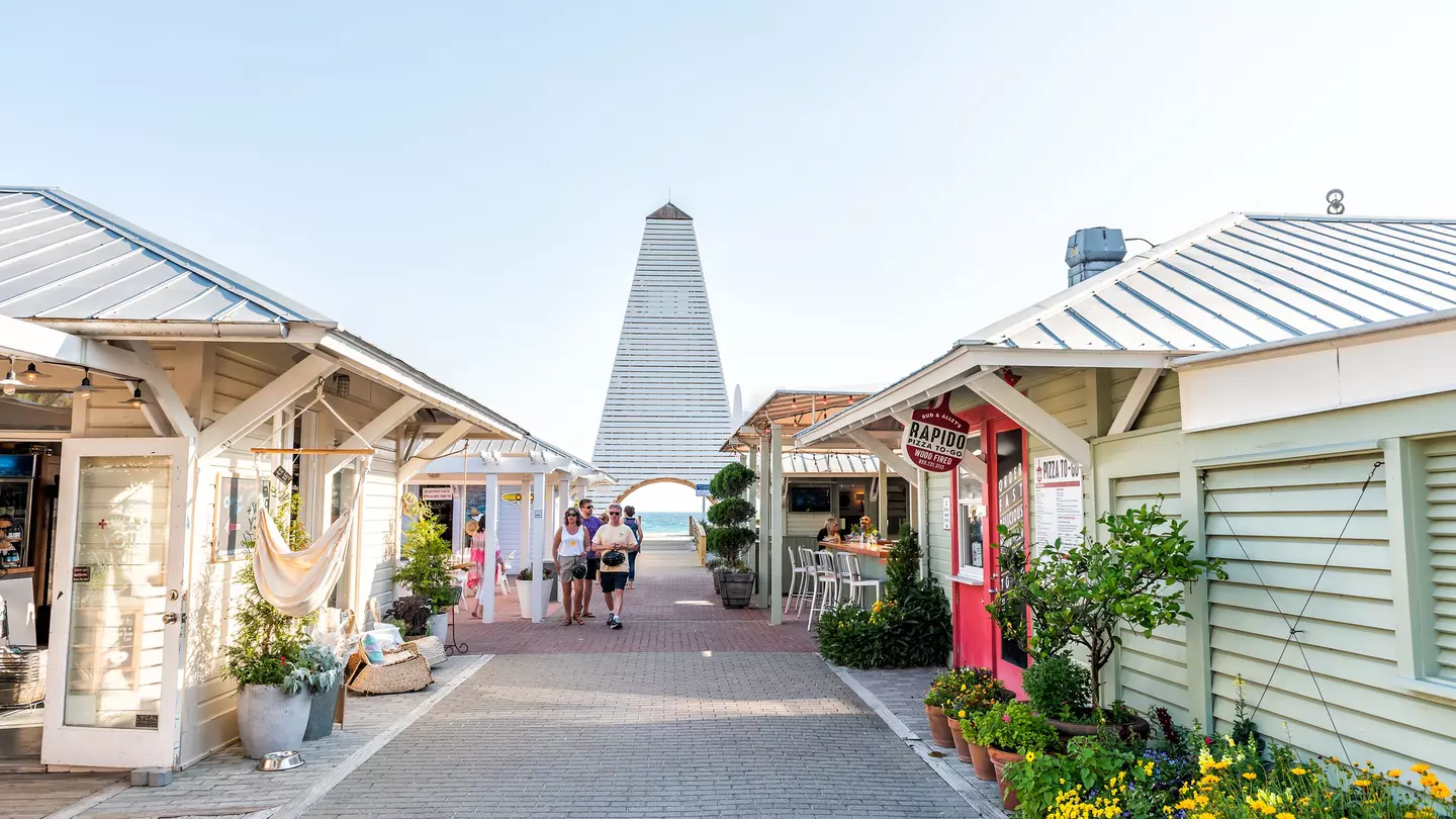 People walk through a pedestrian area in Florida with pastel-colored, one-story shops and cafes; a tower is at the end of the walkway, and the ocean is visible in the opening below.