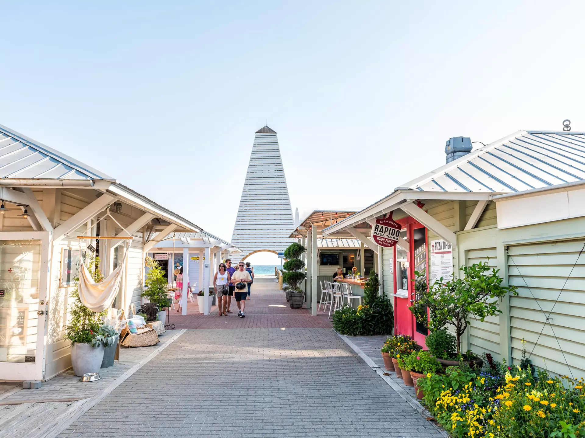 People walk through a pedestrian area in Florida with pastel-colored, one-story shops and cafes; a tower is at the end of the walkway, and the ocean is visible in the opening below.