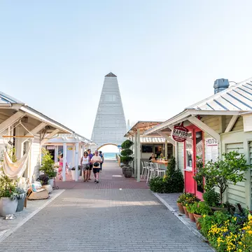 People walk through a pedestrian area in Florida with pastel-colored, one-story shops and cafes; a tower is at the end of the walkway, and the ocean is visible in the opening below.