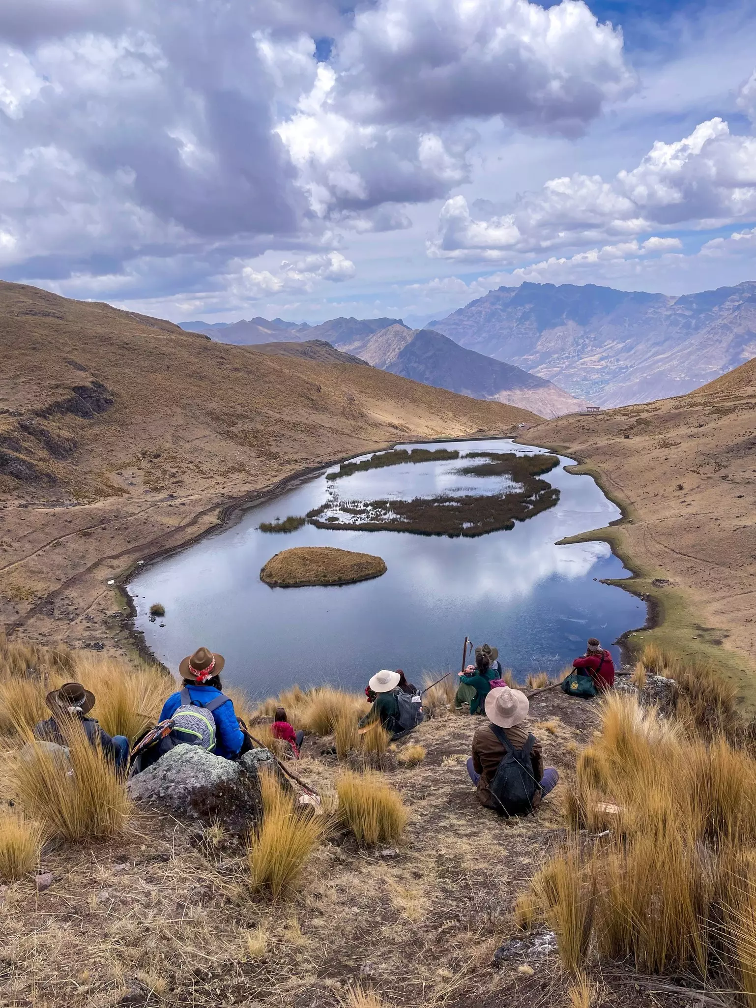 Hikers with their backs to the camera sitting on the ground while looking a lake with mountains in the background