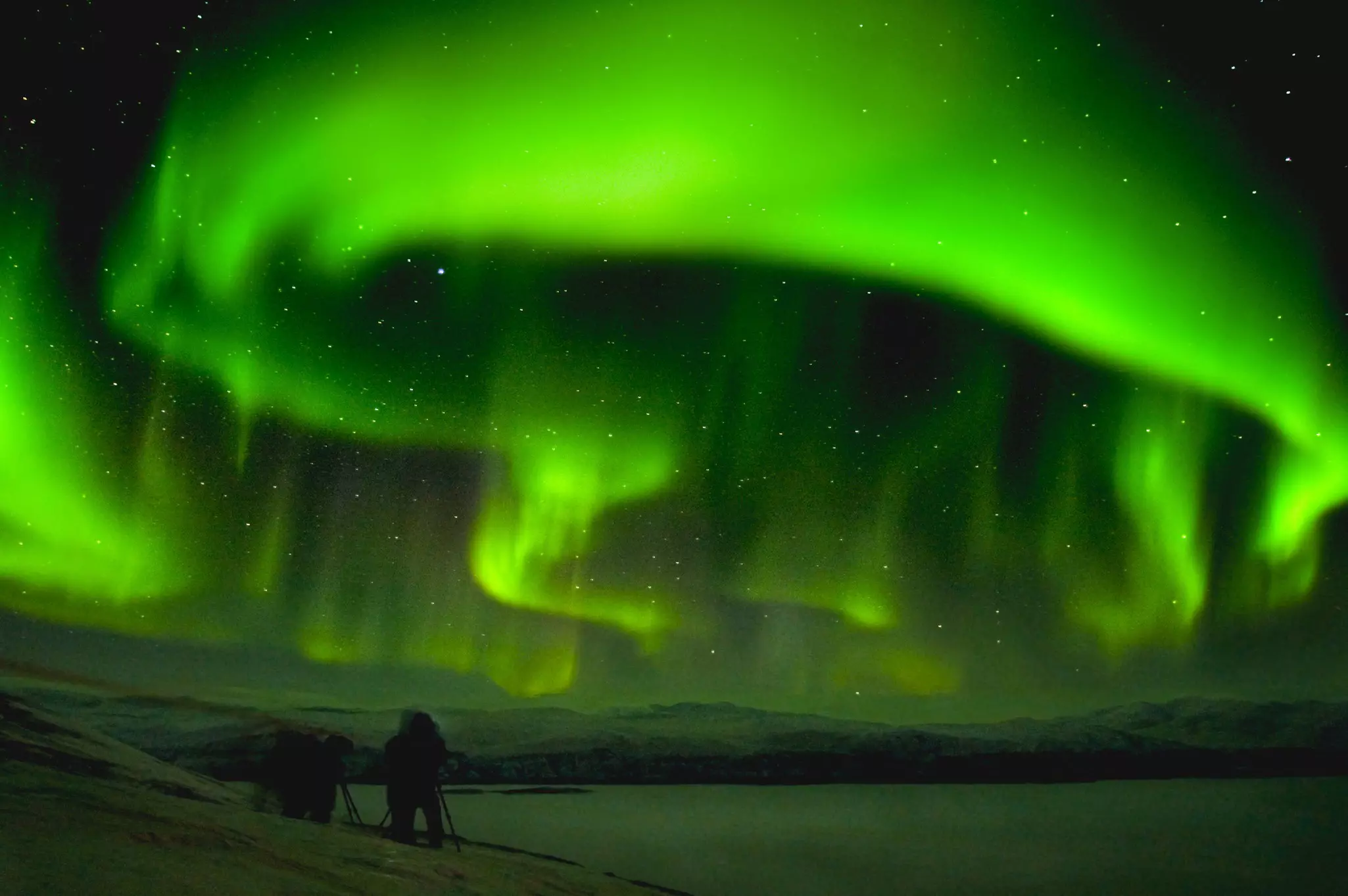 Huge streaks of green northern lights over a dark landscape with the silhouette of a photographer and equipment in the lower left.