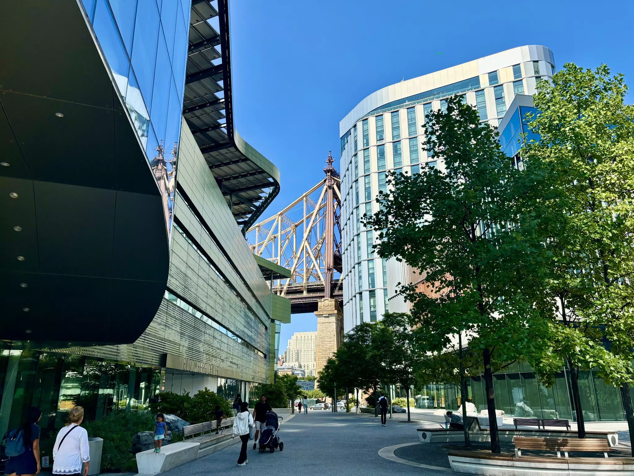 People walk path through the university buildings of the Cornell Tech campus, with a view of the Queensboro Bridge in the distance.