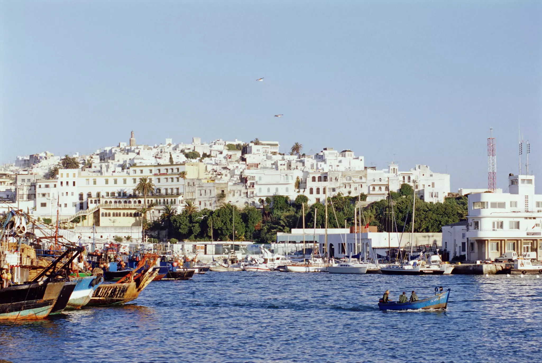 Looking over the waters of the Mediterranean, the white facades of Tangier's old city rises from the shoreline; boats are moored to shore, while a small one makes its way across the harbour.