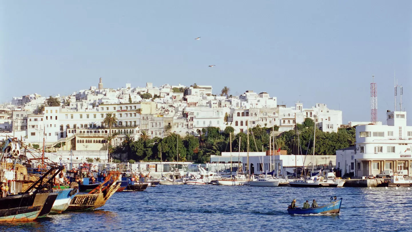 Looking over the waters of the Mediterranean, the white facades of Tangier's old city rises from the shoreline; boats are moored to shore, while a small one makes its way across the harbour.