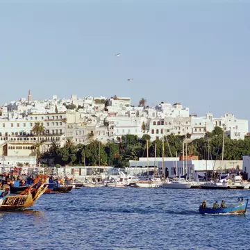 Looking over the waters of the Mediterranean, the white facades of Tangier's old city rises from the shoreline; boats are moored to shore, while a small one makes its way across the harbour.