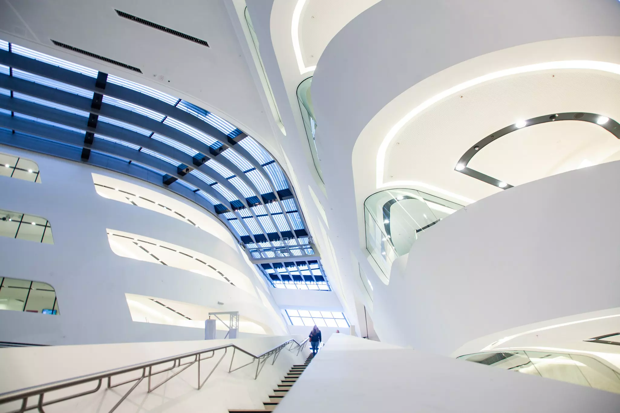 A person descends a flight of stairs within a white atrium with curved walls and a glass ceiling