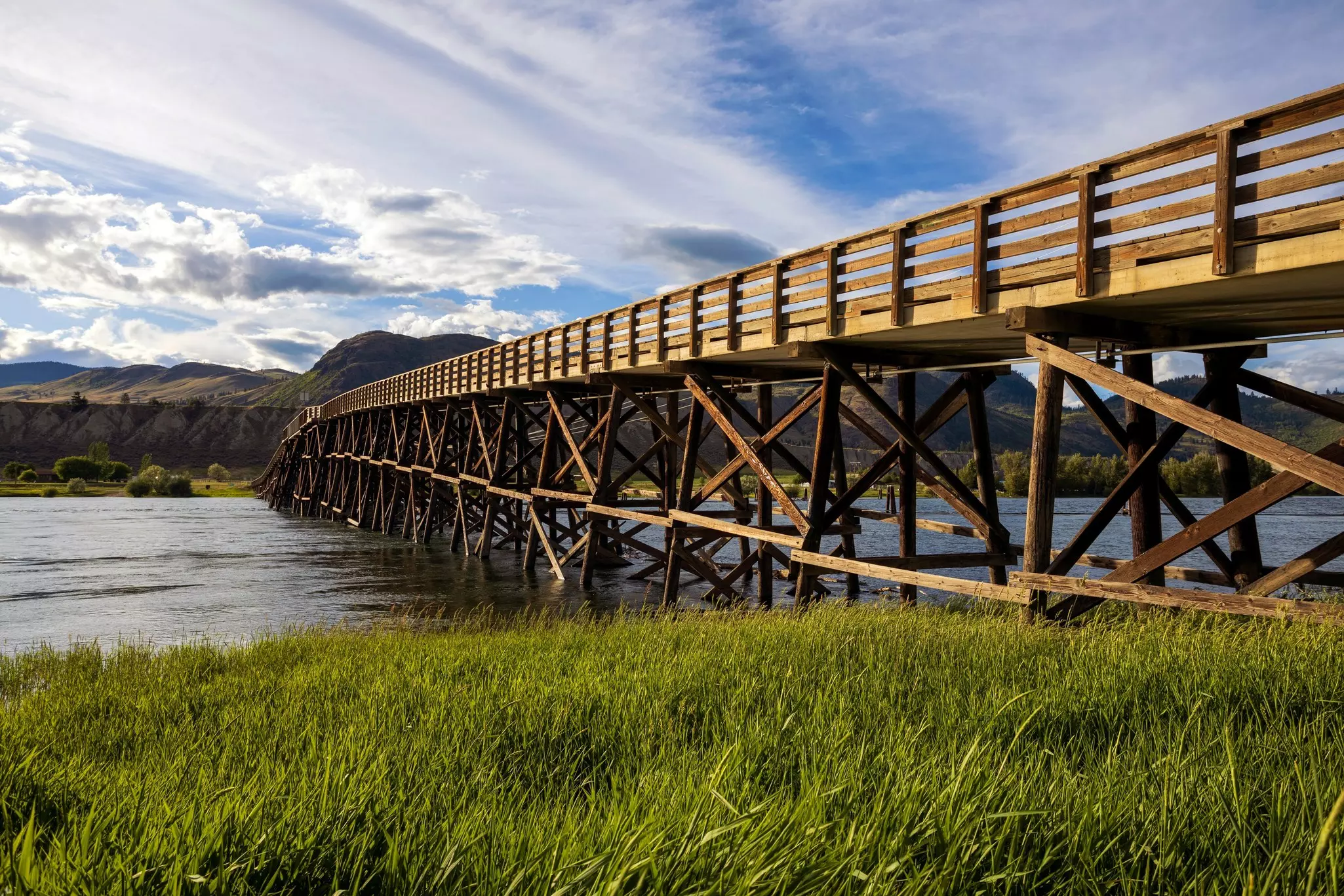 Wooden multi-beam bridge over a wide river with swaying green grass in the foreground and mountains in the distance under a sunny but cloud-filled sky.