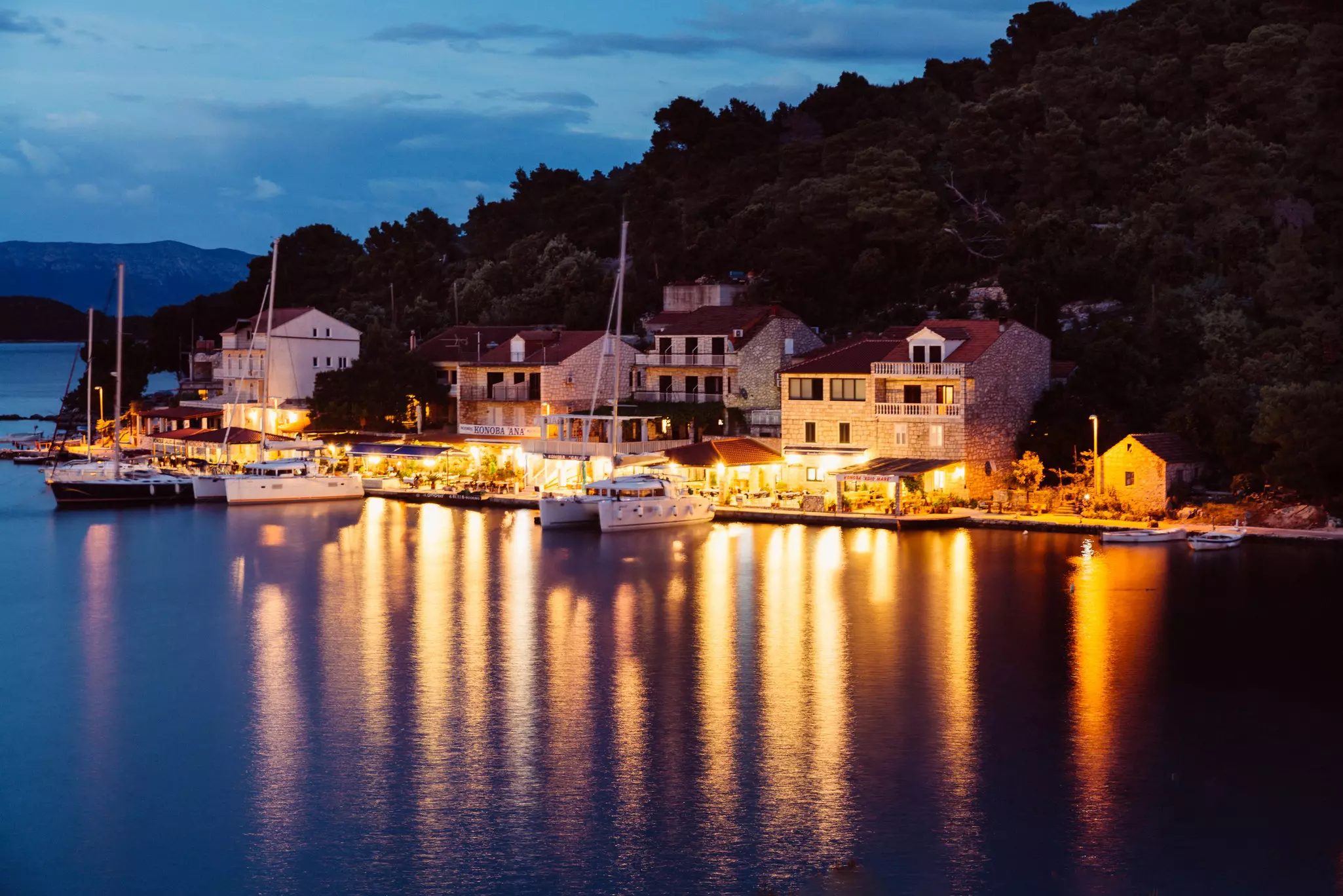 A shoreline lit up at night; there are a few light-colored buildings and boats in the water.