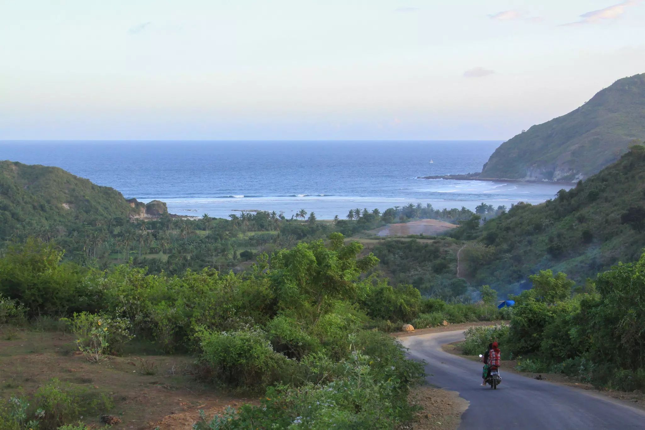 Two people on a scooter ride down a road flanked by jungle towards a beach in Lombok, Indonesia