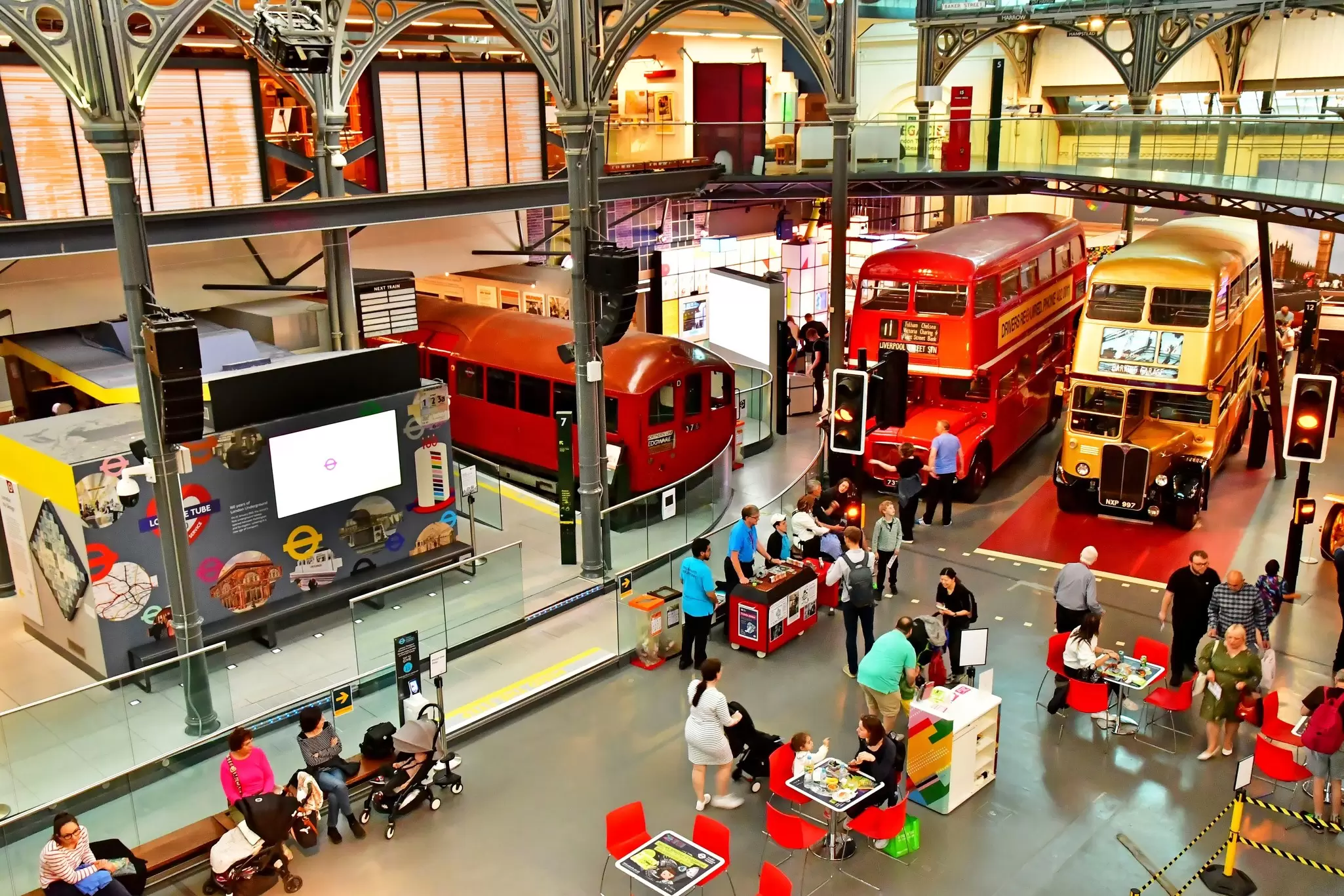 A large exhibition room with double-decker buses and old train carriages. People move around among them.