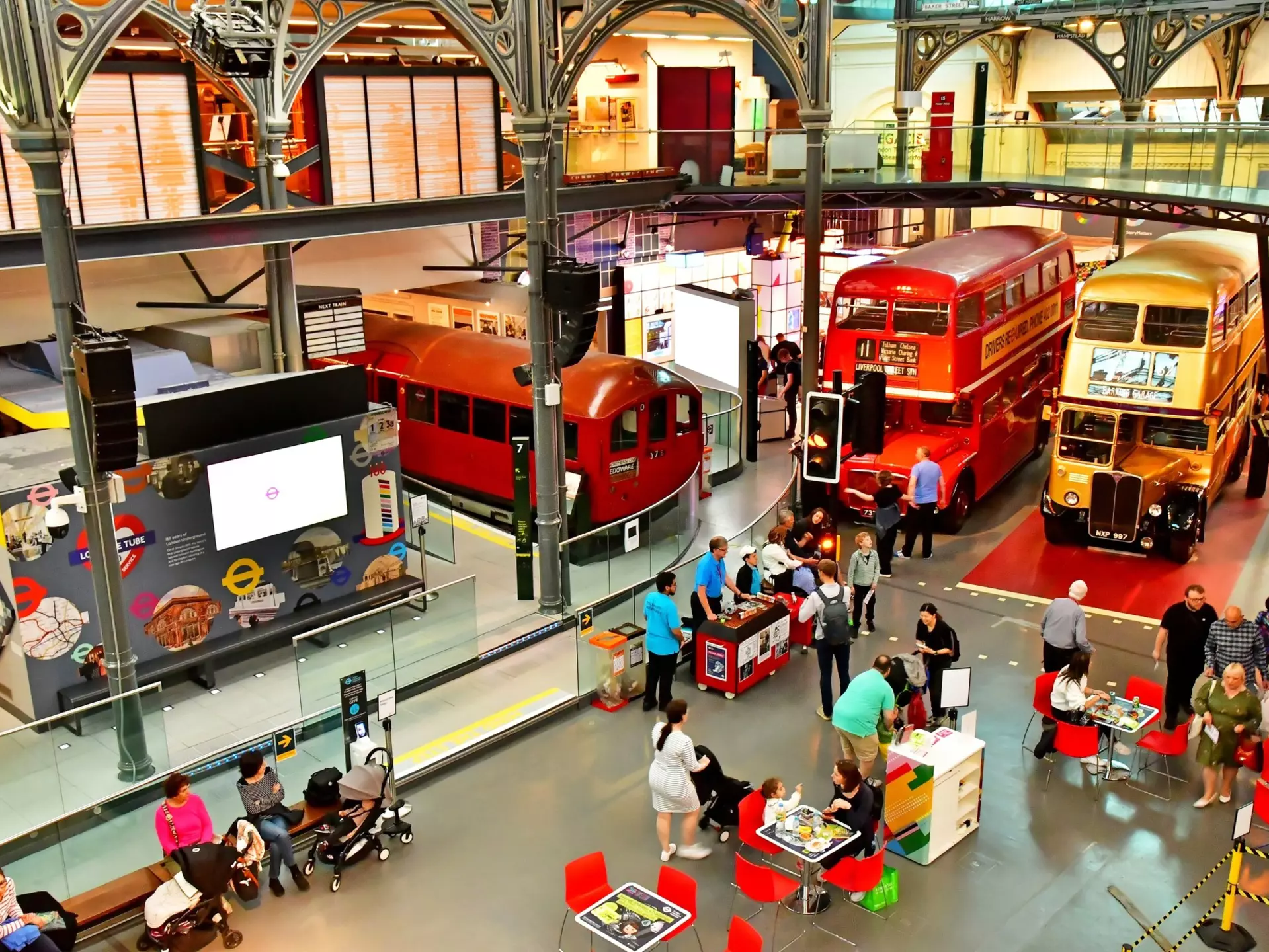 A large exhibition room with double-decker buses and old train carriages. People move around among them.