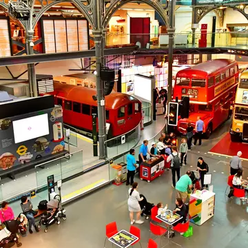 A large exhibition room with double-decker buses and old train carriages. People move around among them.