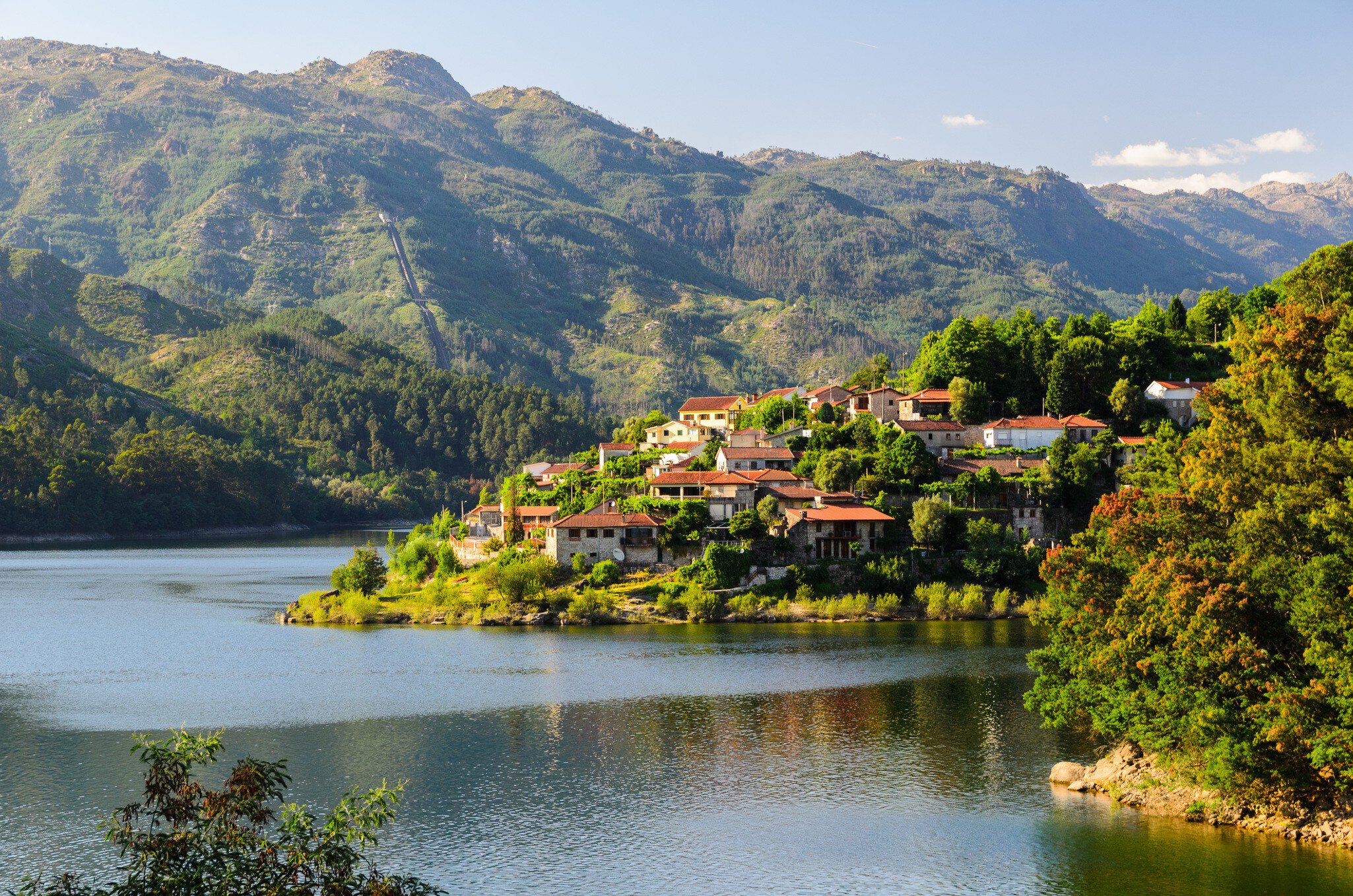 A village clings to a hill rising from the banks of a river with rugged green hills beyond.