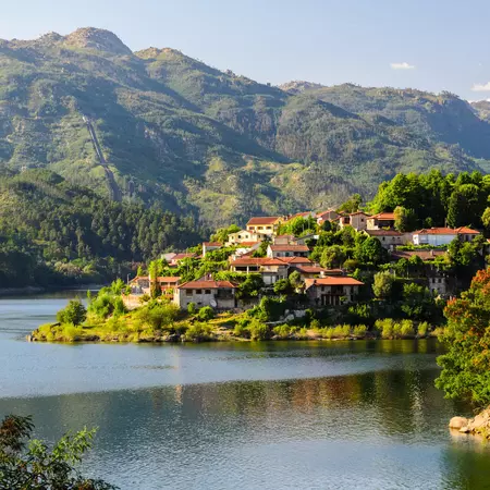 A village clings to a hill rising from the banks of a river with rugged green hills beyond.