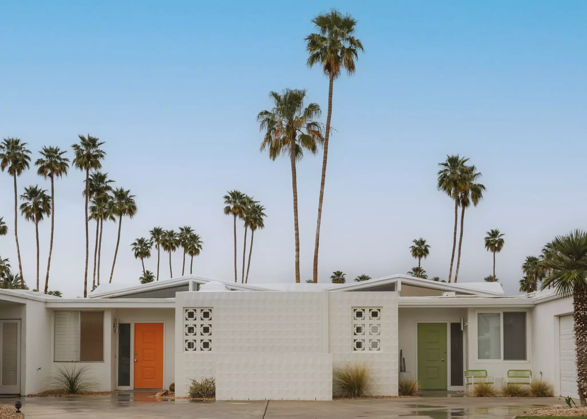 Connected one-story white homes, with an orange door on one side and a green door on the other; tall palm trees are behind the building.