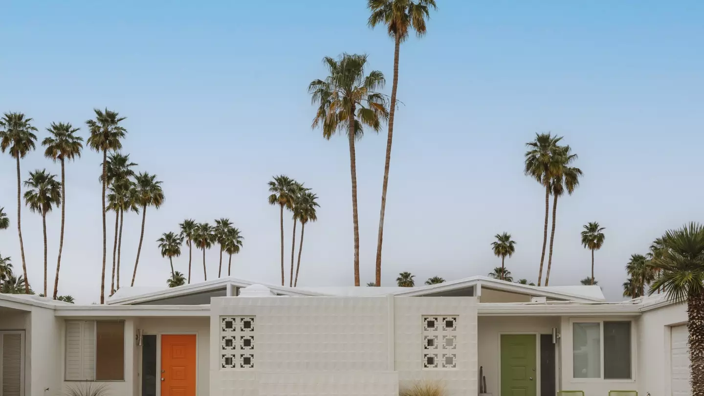 Connected one-story white homes, with an orange door on one side and a green door on the other; tall palm trees are behind the building.