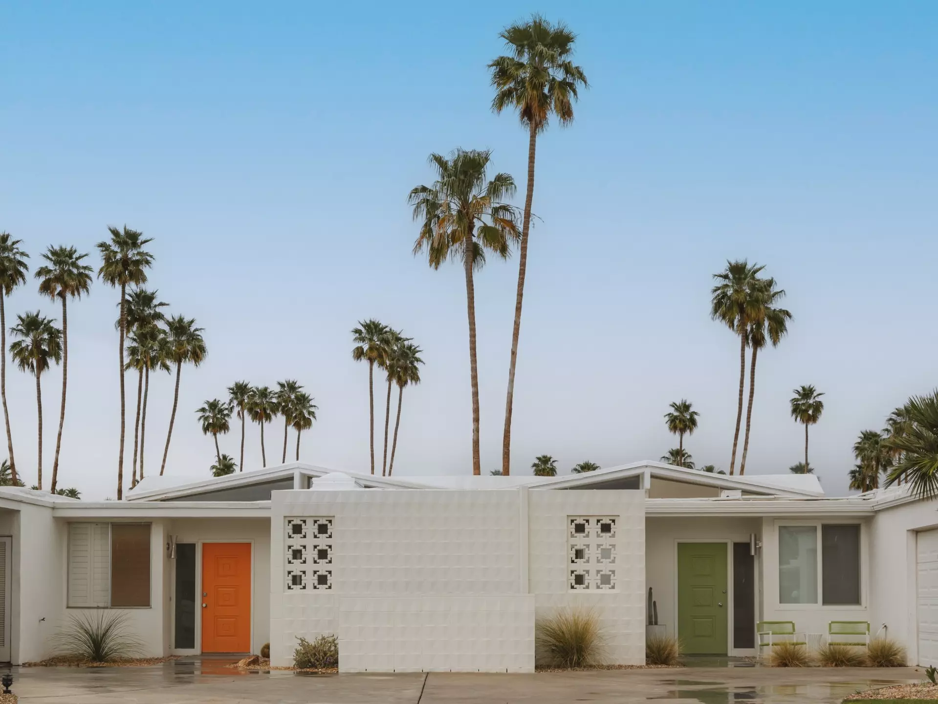 Connected one-story white homes, with an orange door on one side and a green door on the other; tall palm trees are behind the building.