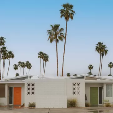 Connected one-story white homes, with an orange door on one side and a green door on the other; tall palm trees are behind the building.